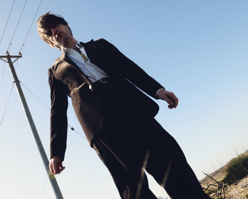 Low-angle shot of a man in a black suit and striped shirt standing in an outdoor setting with an electrical pole in the background.