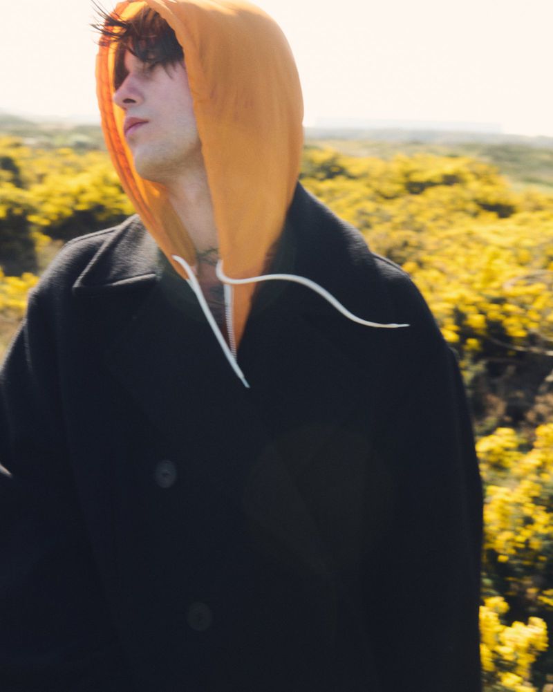 Adult male stands in field of yellow flowers wearing a black overcoat and orange hoodie on a bright day in nature.