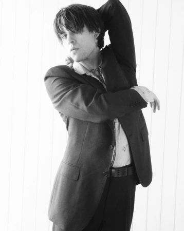 Portrait of a young man in a suit against a white paneled wall. The monochrome image captures a contemplative pose with a relaxed expression.