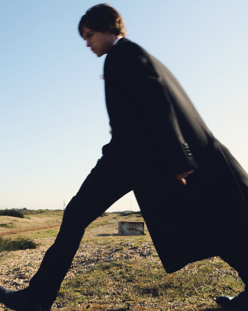 Young adult in black coat strides across field under open sky. Minimalist fashion shot with horizon line and natural lighting.