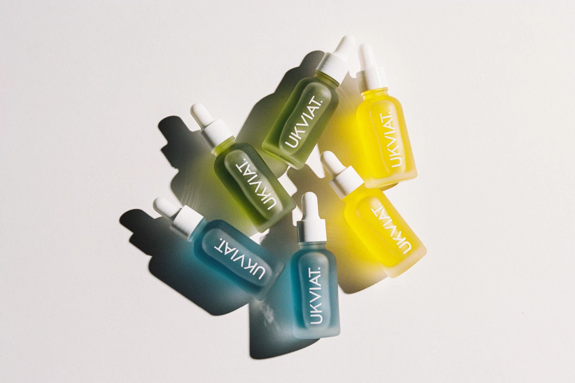 Overhead shot of Ukyiat beauty serums in blue, green, and yellow bottles with white dropper caps arranged on a white surface.