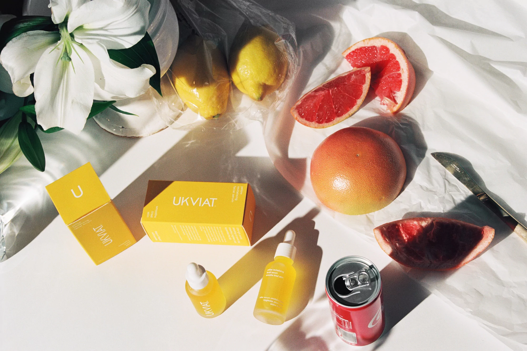 Still life of U K VIAT beauty products, citrus fruits, and a soda can on crinkled white fabric in bright, natural lighting.