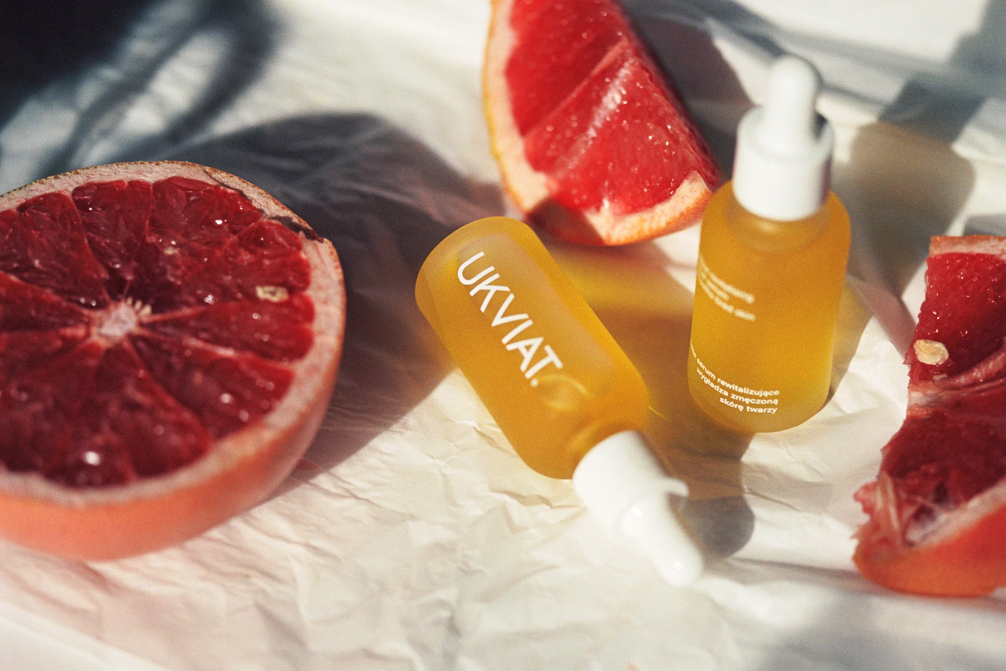 Two bottles of yellow face serum with dropper caps are displayed with slices of red grapefruit on white crumpled paper.