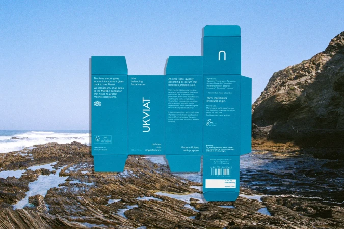 Blue packaging for UKVIAT skincare stands against a backdrop of rocky coastline and ocean waves. A product shot for skincare.