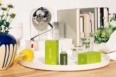 Green skincare serum bottle and boxes on a white tray with books, glasses of water, flowers and a lemon.