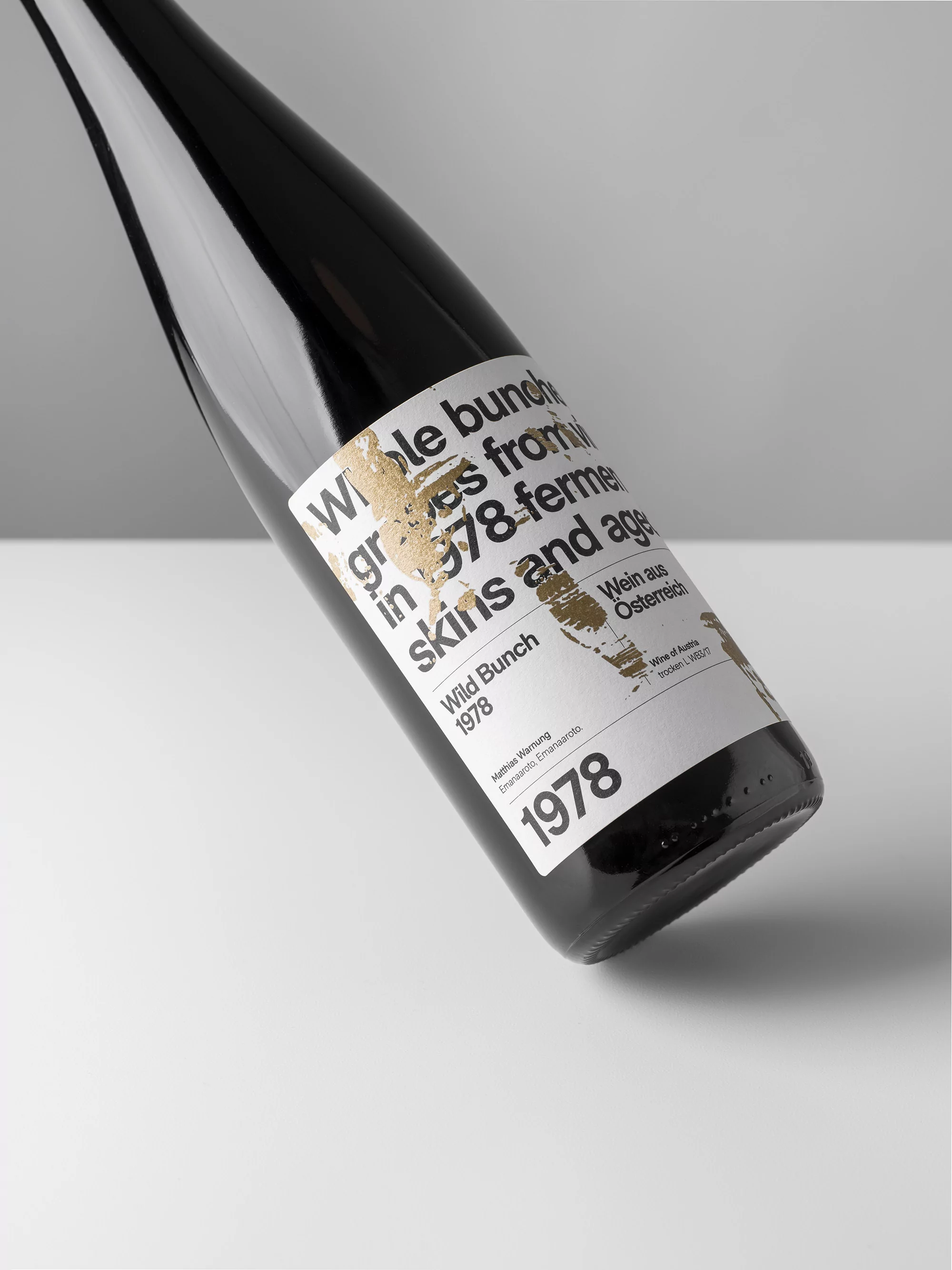 Close-up studio shot of a dark glass wine bottle with a white label and gold-colored text on a white surface. Wine product shot.