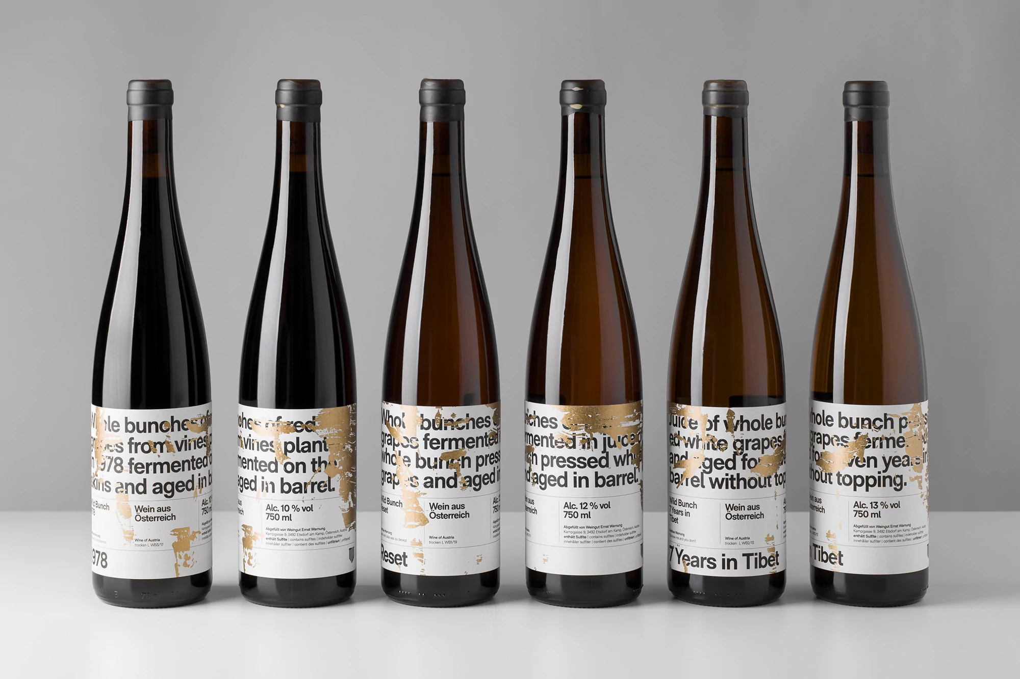 Six wine bottles stand in a row, each with labels displaying unique text and design elements against a neutral background.