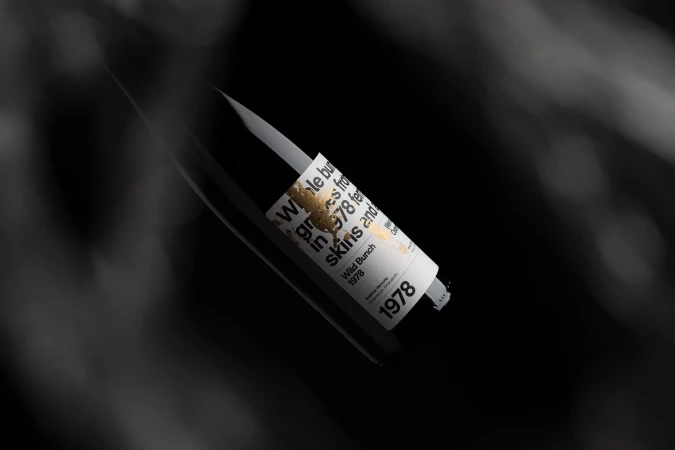Angled shot of a black wine bottle with a white label featuring gold accents and black text, set against a dark, blurred background.