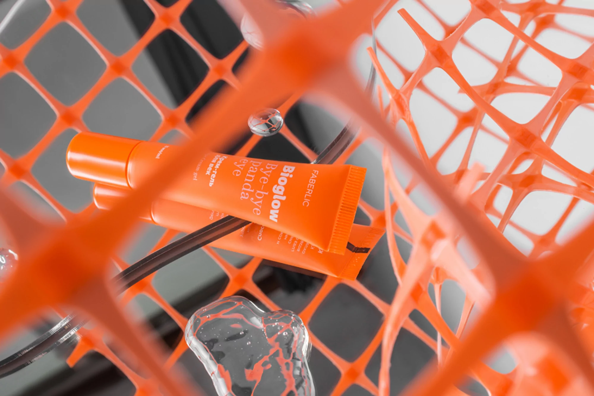 Two tubes of Faberlic Bioglow Bye-Bye Panda Eye cream sit on a reflective surface with an orange net and water droplets.
