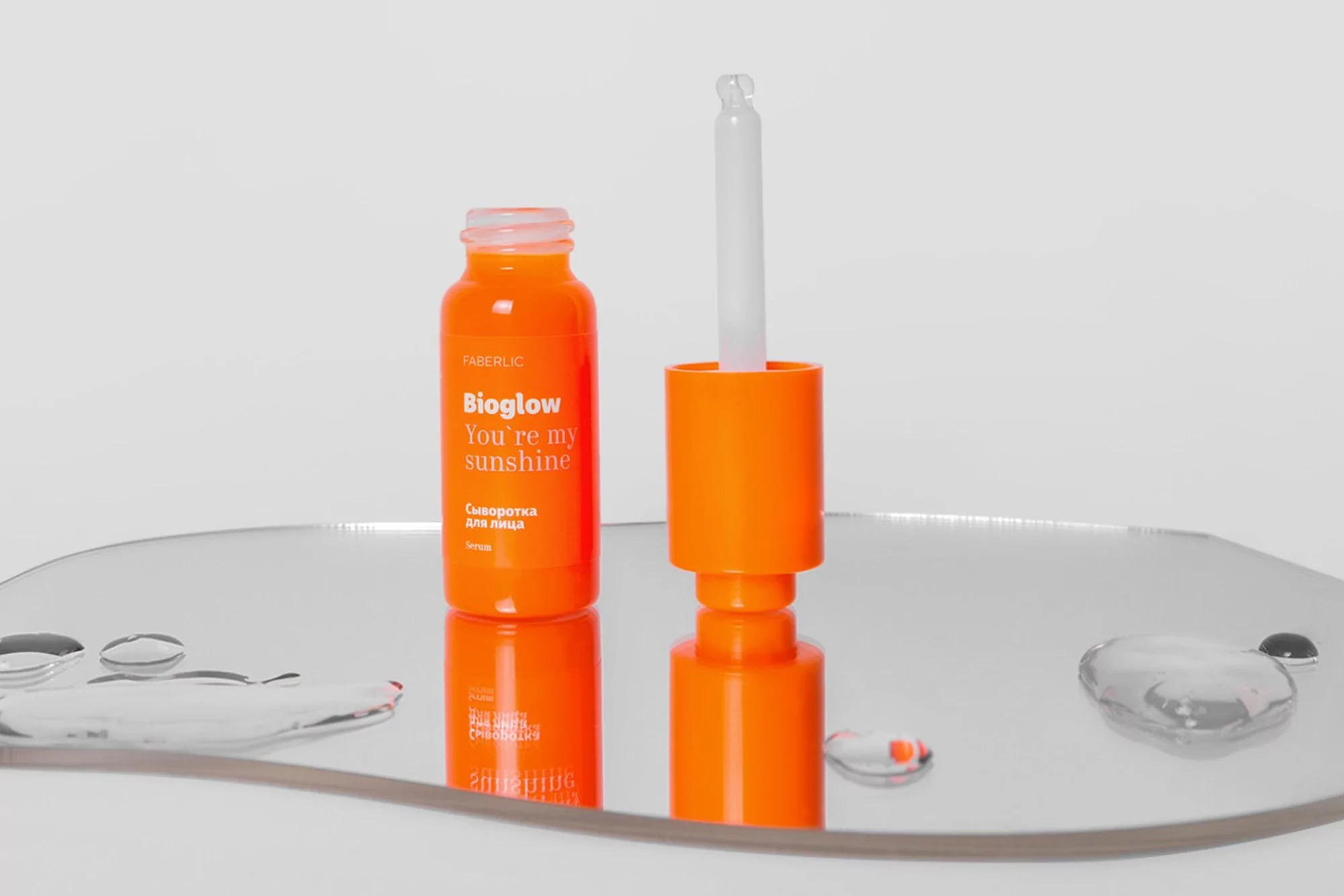 Orange Bioglow facial serum bottle with dropper on a reflective surface against a white background in a beauty still life.