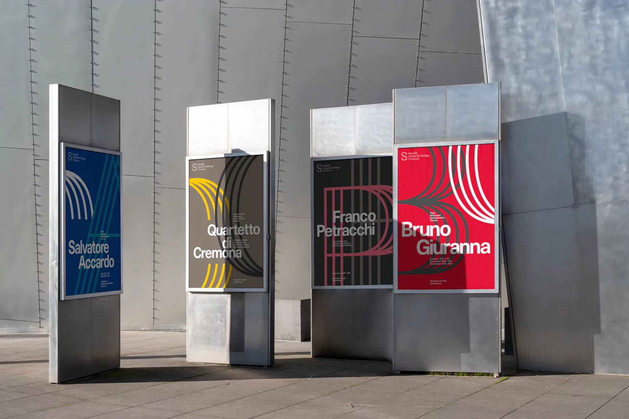 Four tall metallic pillars displaying graphic event posters with typography set outdoors against a modern curved building exterior.