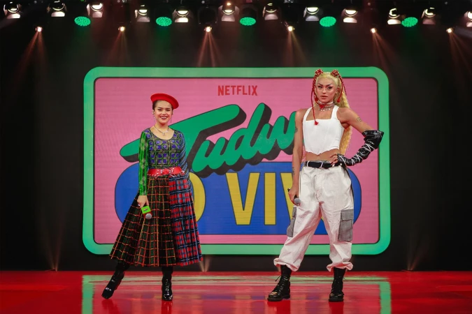 Two people stand on stage with a Netflix Tudum ao Vivo backdrop. One is wearing a red beret and colorful dress, the other is wearing a white top.