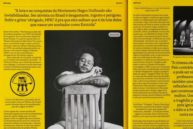 Black and white photo of Emicida with his eyes closed, sitting in a chair with newspaper clippings surrounding him.