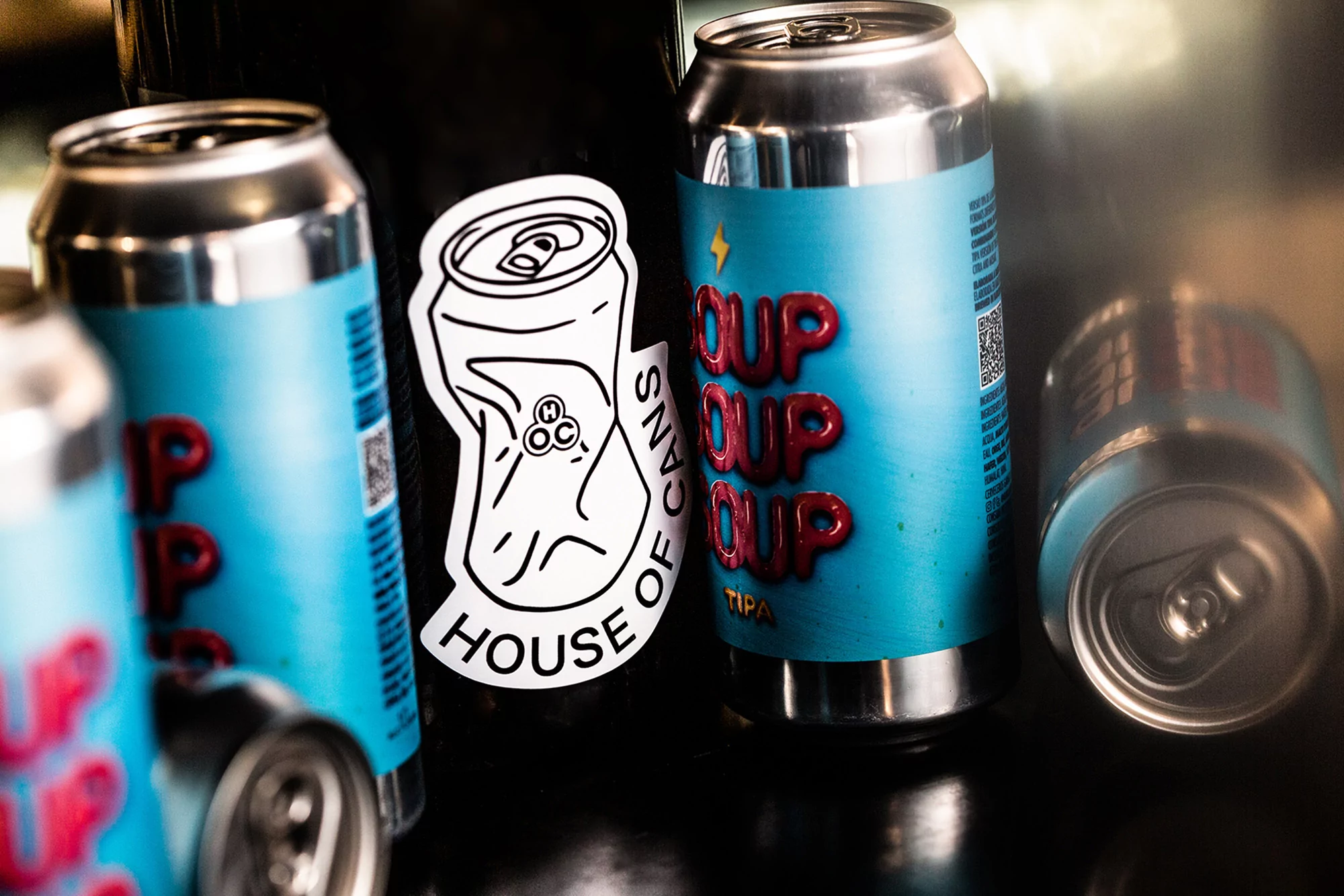 Multiple aluminum cans with light blue labels and red text, stacked with a 'House of Cans' sticker on a black, reflective surface.