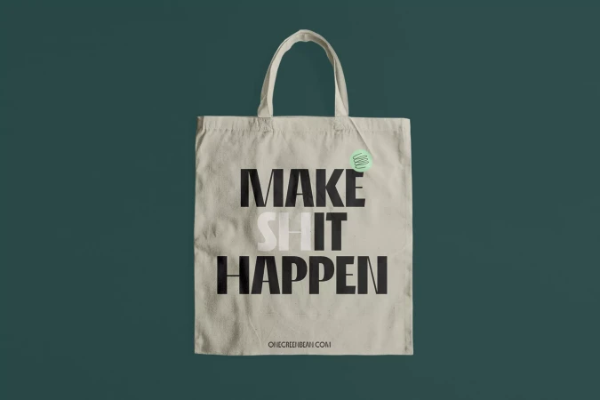 A cream canvas tote bag with the text "MAKE SHIT HAPPEN" displayed against a solid dark teal background.