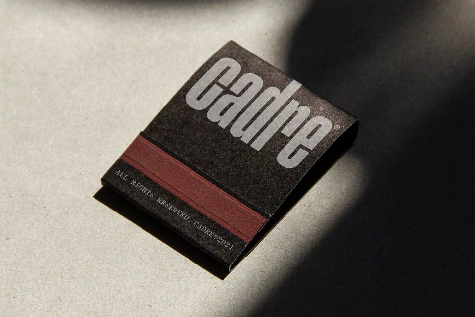 Dark grey matchbook with a rust-colored striking strip and a white sans-serif logo sits on a light grey surface with a strong shadow.