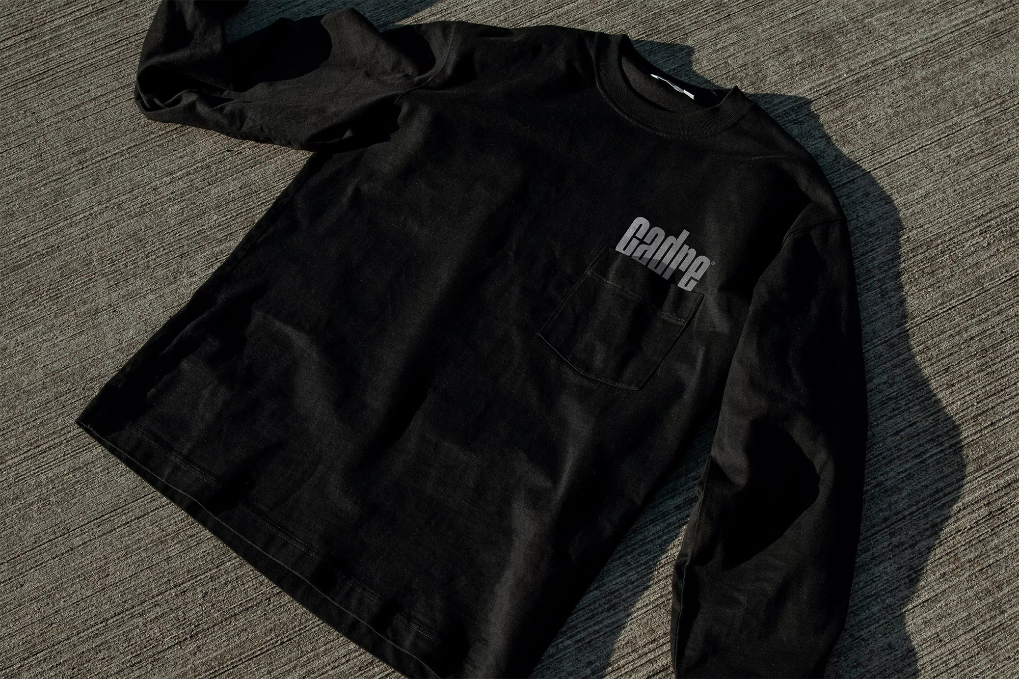 Close-up of a black, long-sleeved t-shirt with the text "Cadre" on the pocket, laid on a textured concrete surface.