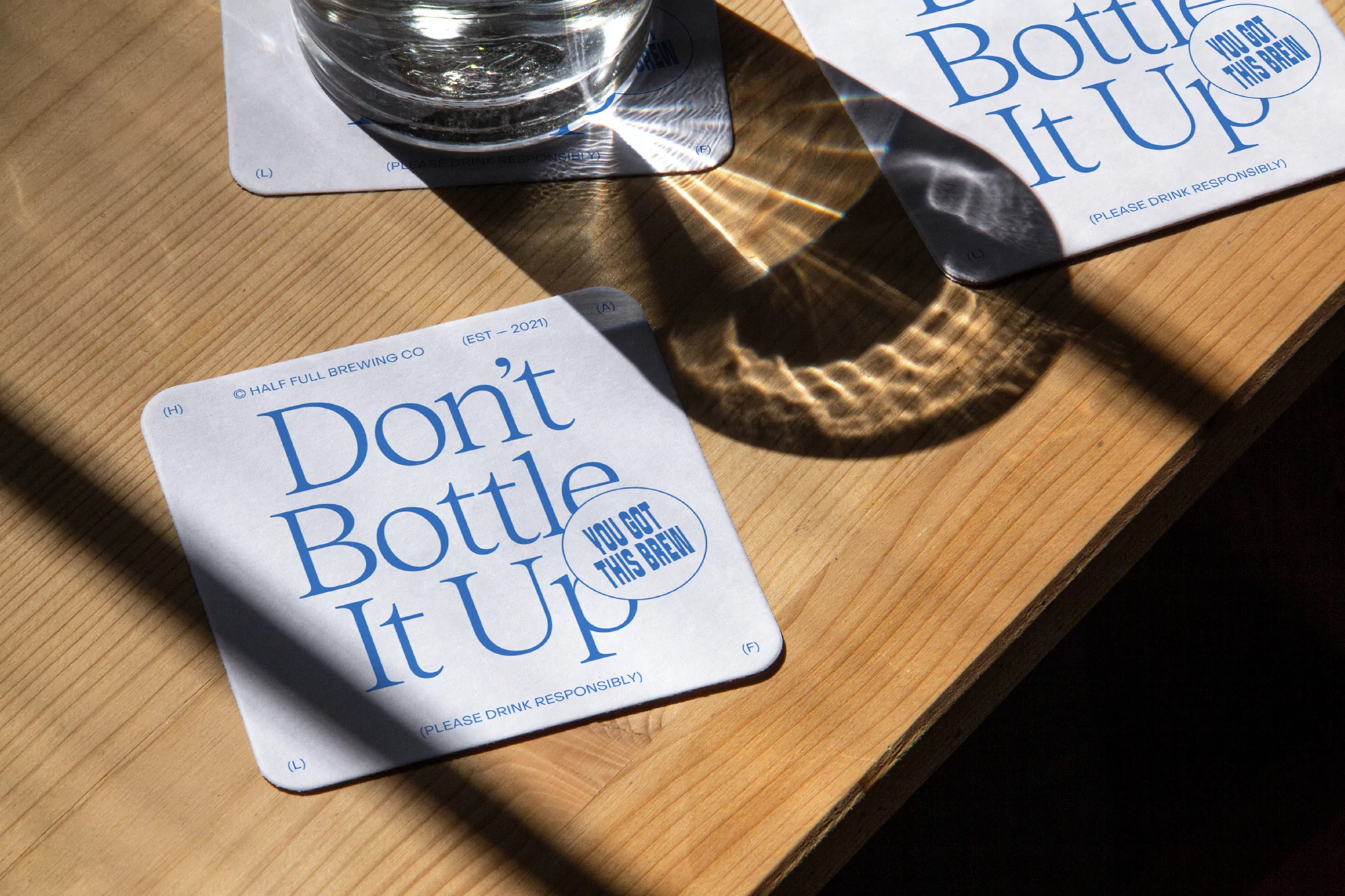 Close-up of a Half Full Brewing Co. beer coaster and water glass on a light brown wooden table with sunlight creating shadows.