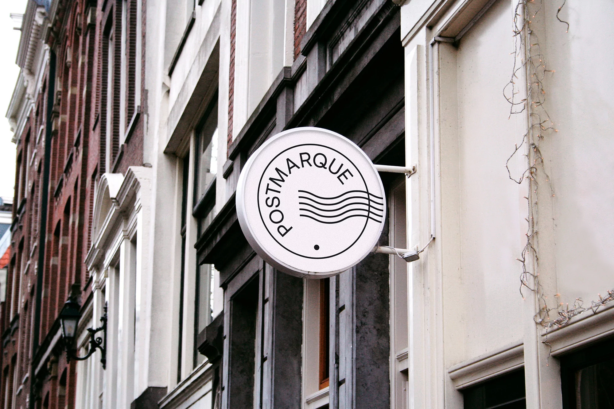 Illuminated round sign for 'Postmarque' hangs on the exterior of a building with brick and off-white facade in an urban environment.