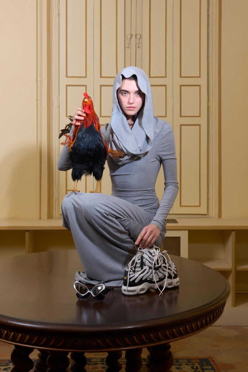 Person in hooded gray dress squats on table holding rooster and sneakers in an indoor setting with neutral colored walls.