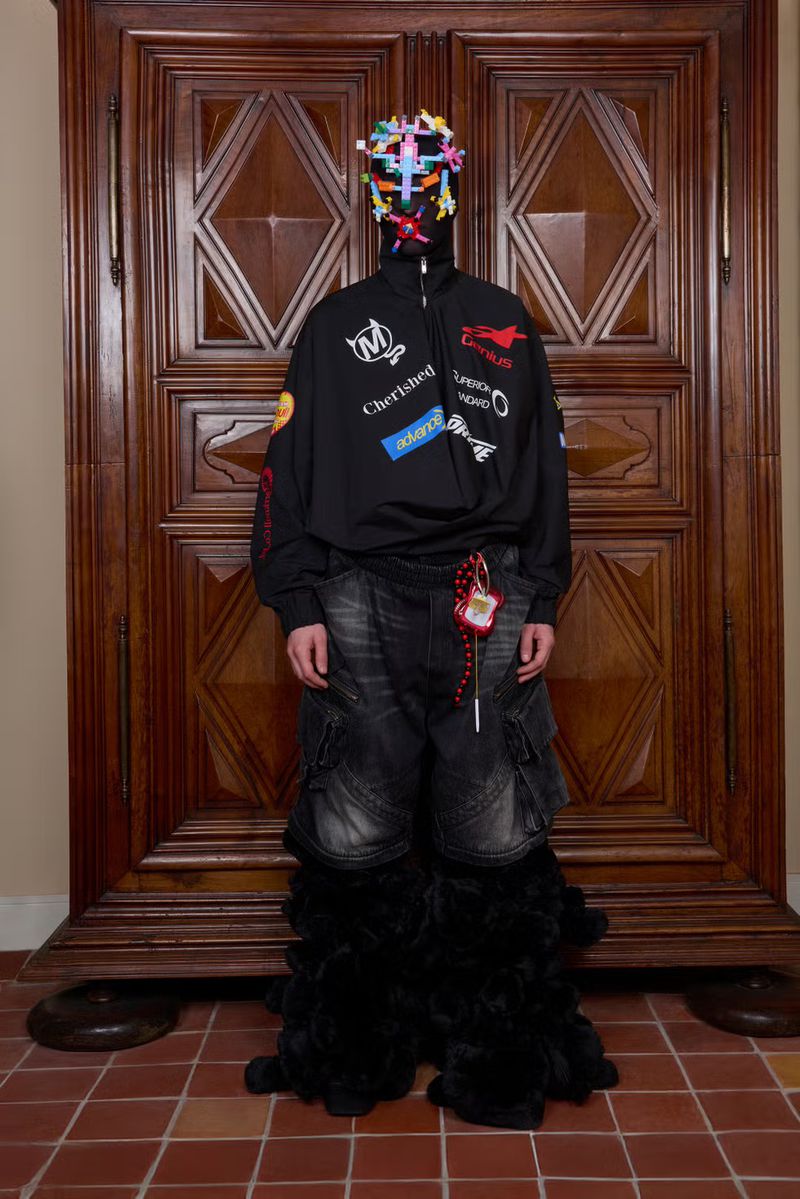 Person wearing black jacket with patches, denim pants with fur trim, and a multicolored mask stands in front of wooden doors.