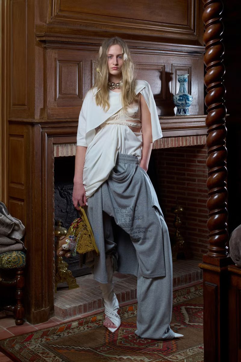 Fashion shot of a woman in an asymmetrical white top and grey pants, standing in front of a fireplace in an indoor setting.