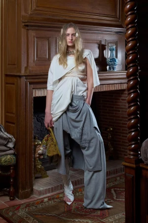 Fashion shot of a woman in an asymmetrical white top and grey pants, standing in front of a fireplace in an indoor setting.