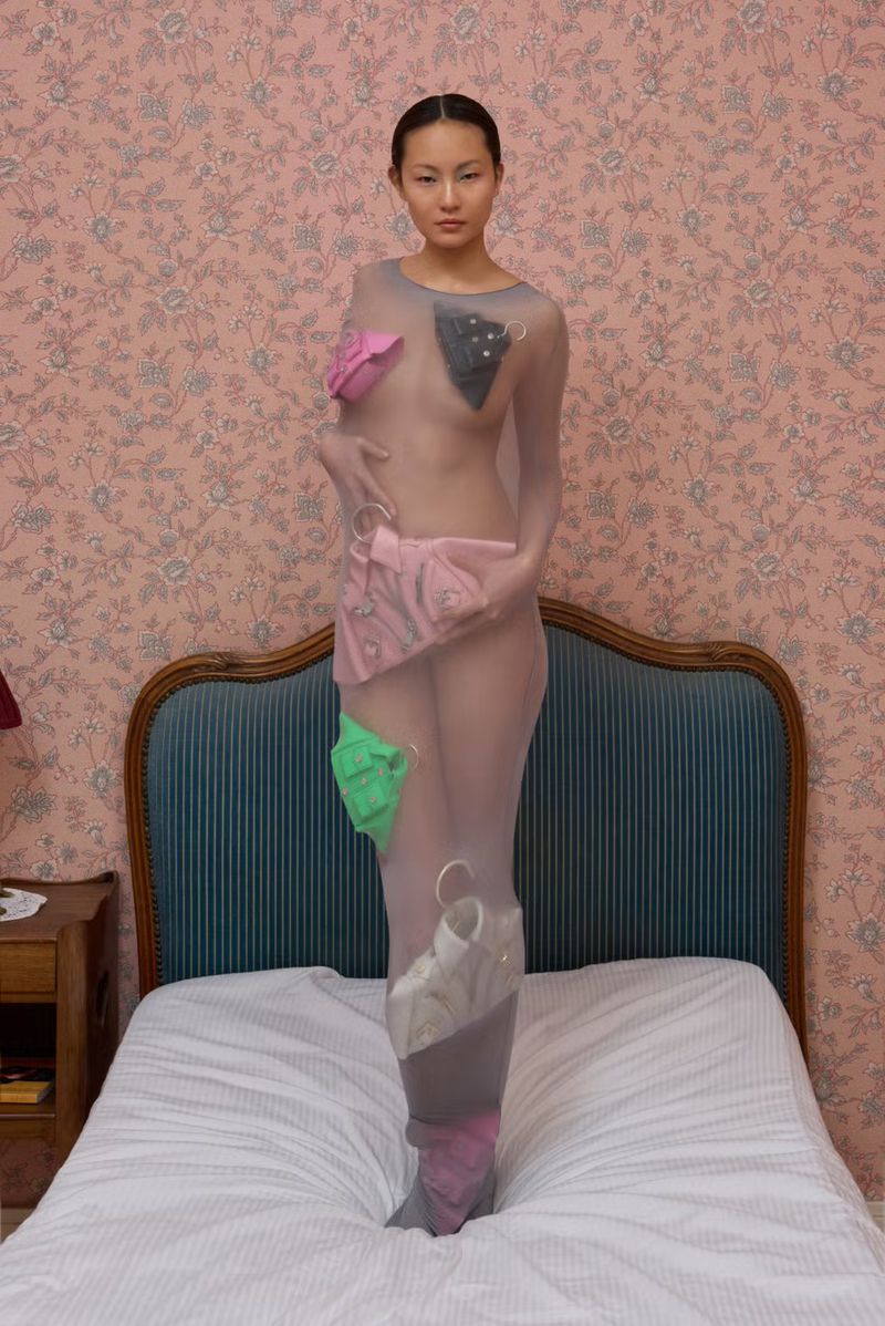 Artistic full shot of a slender Asian woman covered in sheer fabric on a bed with pink wallpaper. The outfit has colorful fabric pieces attached.