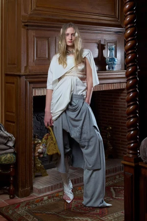 Model standing in front of a carved wooden fireplace wearing a white draped top and loose grey trousers