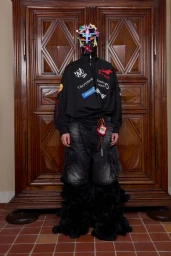 Model wearing a black zip top, dark cargo shorts, and a colourful building block face mask standing before wooden doors
