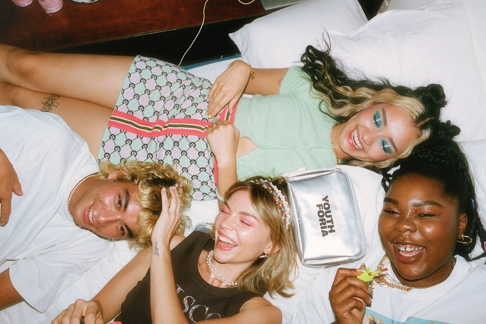 Four smiling young adults lying on a white bed in casual clothing. They face up towards the camera, creating a bright and cheerful scene.