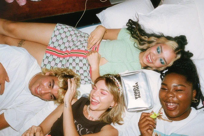Four smiling young adults lying on a white bed in casual clothing. They face up towards the camera, creating a bright and cheerful scene.