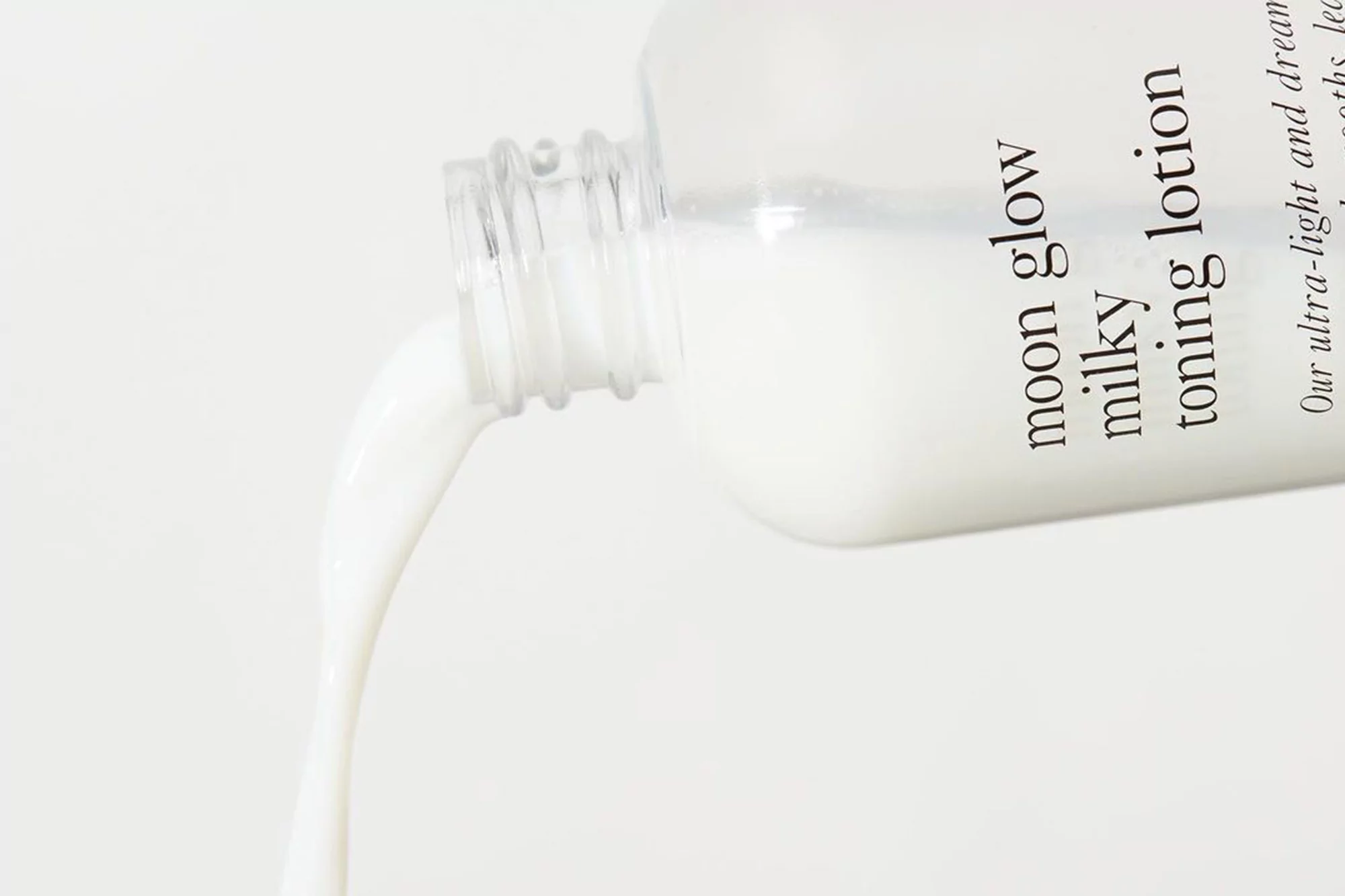 Close-up of white lotion being poured from a clear bottle, with the product name displayed on the side against a light backdrop.