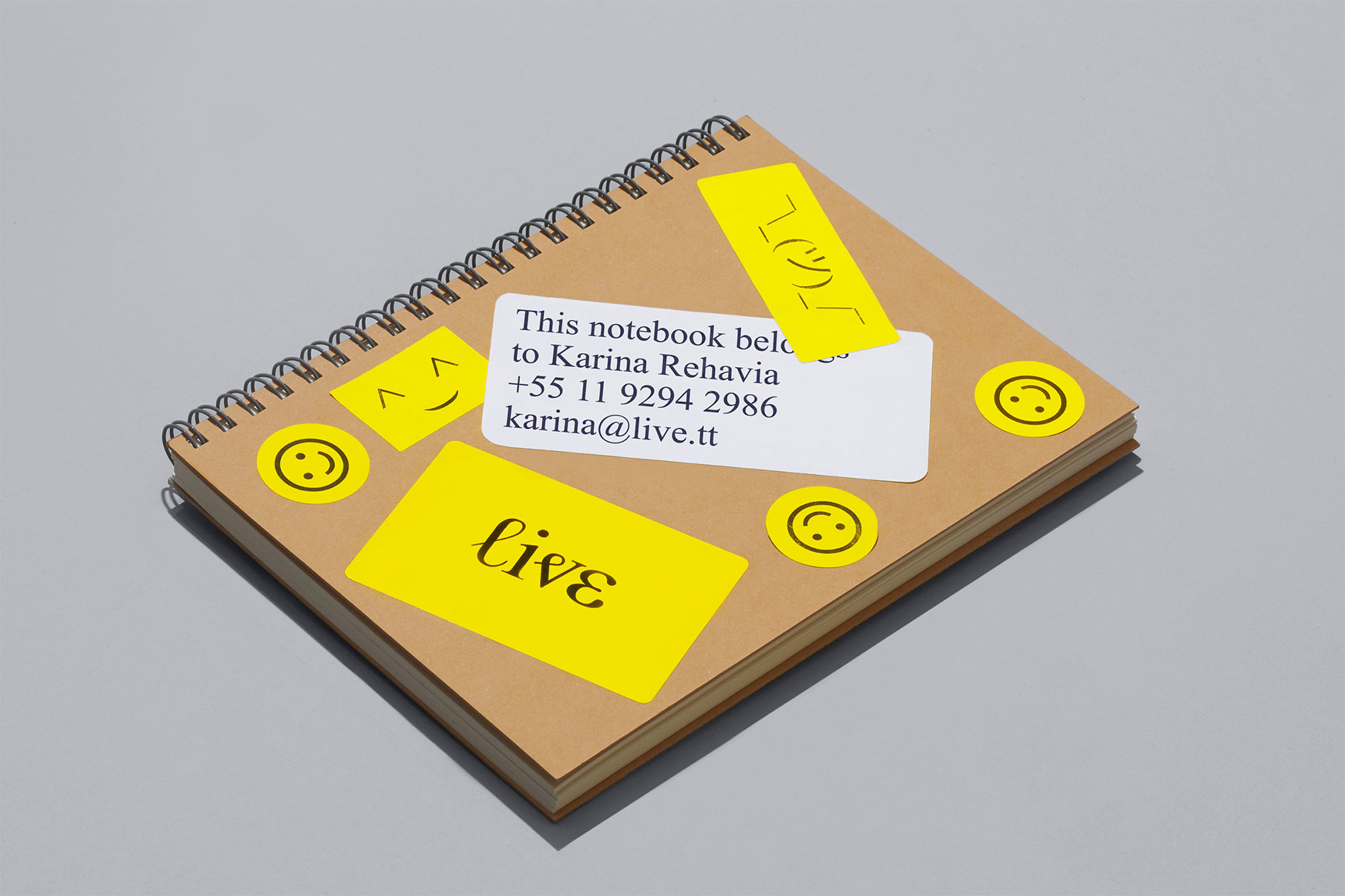 Overhead shot of a spiral-bound notebook with yellow smiley face stickers and contact info on a light gray background, neutral lighting.