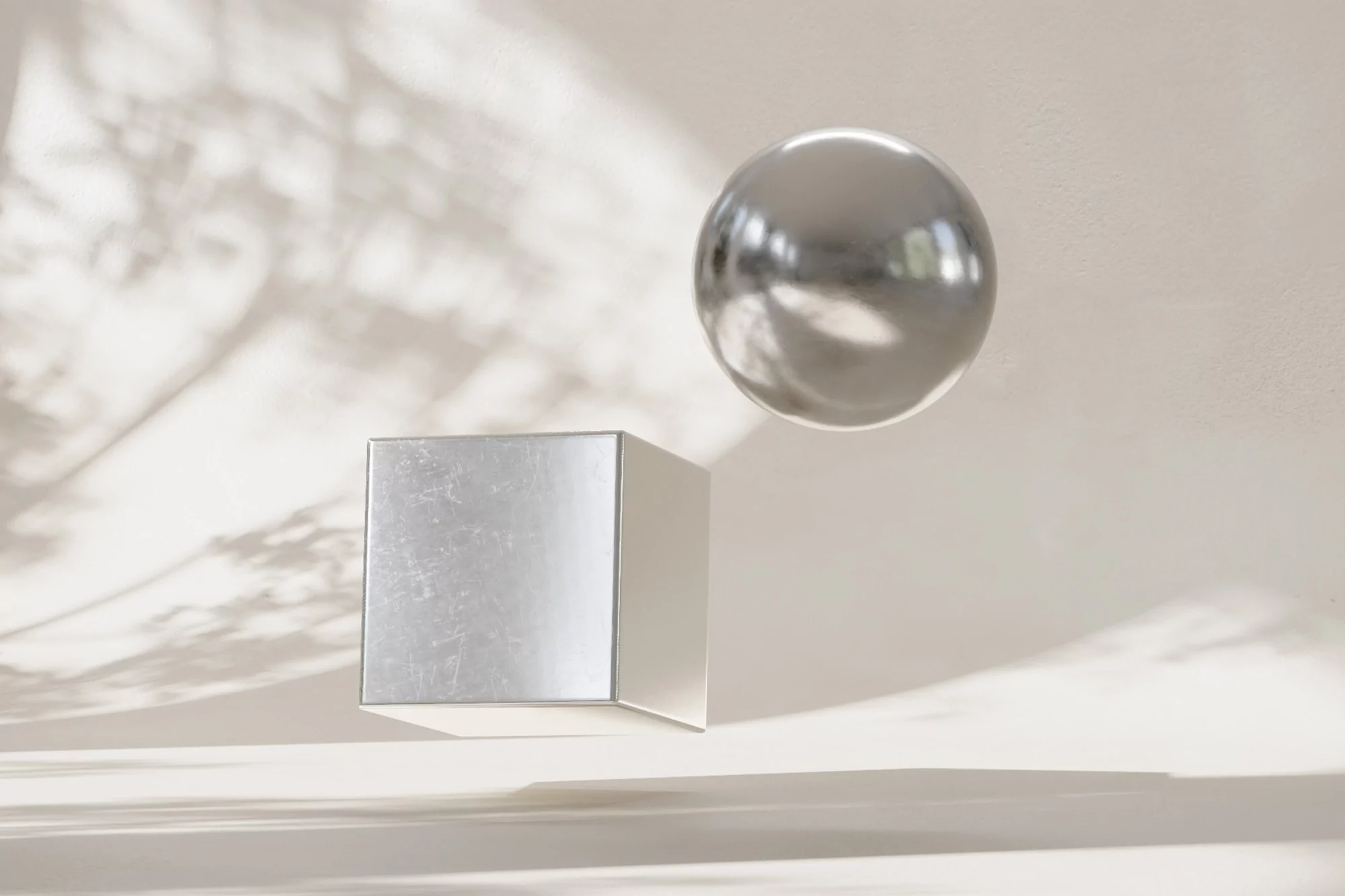 Abstract geometric composition featuring a silver cube and sphere under soft light with tree branch shadows on a neutral beige backdrop.