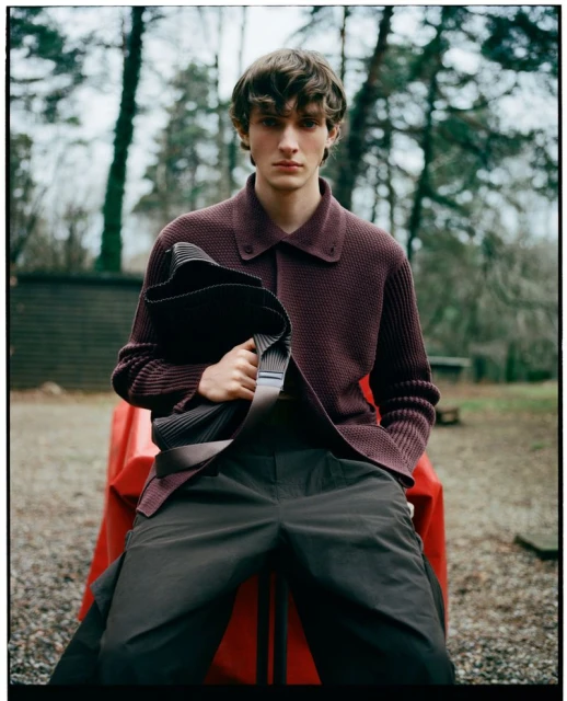 Young man sits on a red surface outdoors, wearing a plum knit shirt, dark pants, and holding a black folded item, with trees in background.