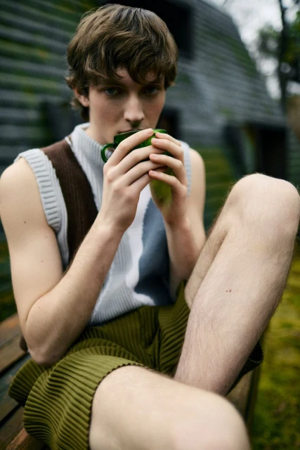 A young man sits outdoors drinking from a green mug, wearing a sleeveless shirt and shorts in an outdoor setting with green foliage.