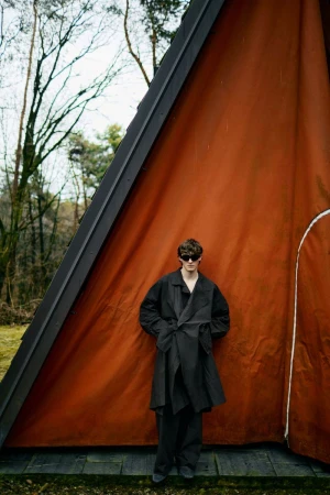 A lone adult stands near a striking orange A-frame cabin in a forest setting, wearing a dark coat and sunglasses, creating a sense of mystery.