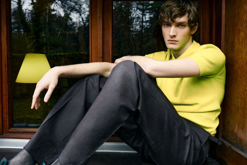Young man in yellow sweater and grey trousers sits in a window with a lamp, reflecting greenery outdoors, looking directly at camera.