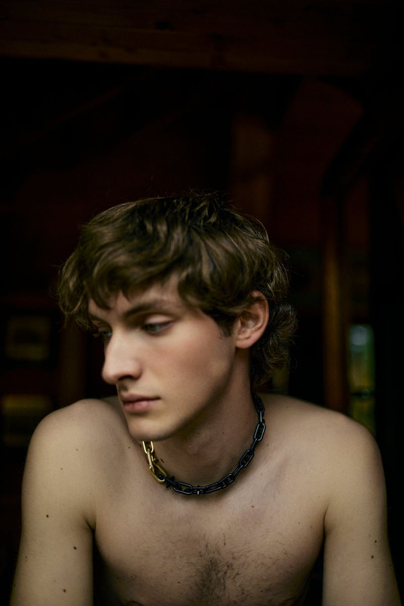 Portrait of a young, shirtless adult male with curly brown hair, wearing a silver and gold chain necklace, looking down in a dark indoor space.