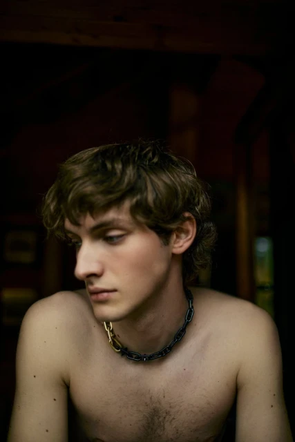 Portrait of a young, shirtless adult male with curly brown hair, wearing a silver and gold chain necklace, looking down in a dark indoor space.