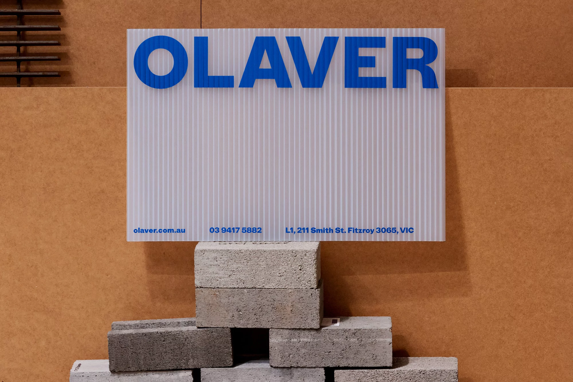 Signage featuring the name 'OLAVER' in blue lettering on a white corrugated background, displayed on stacked concrete blocks indoors.