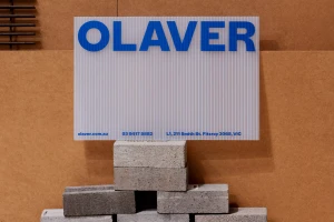 OLAVER Architecture brand identity sign on corrugated translucent panel resting on concrete blocks against brown panels