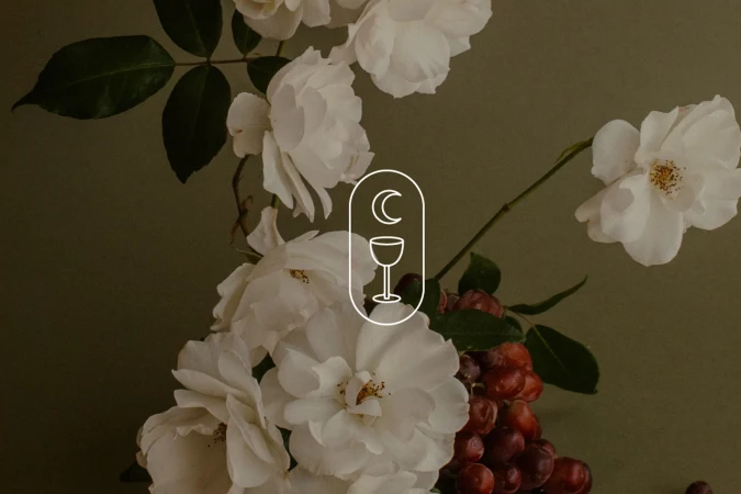 White roses and red grapes against a muted dark background with a central white outline logo featuring a moon and goblet.