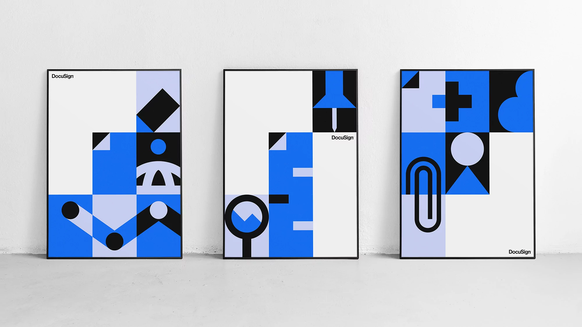 Three framed posters display geometric designs with squares, circles, triangles, and lines in blue, black, and white against a gray wall.