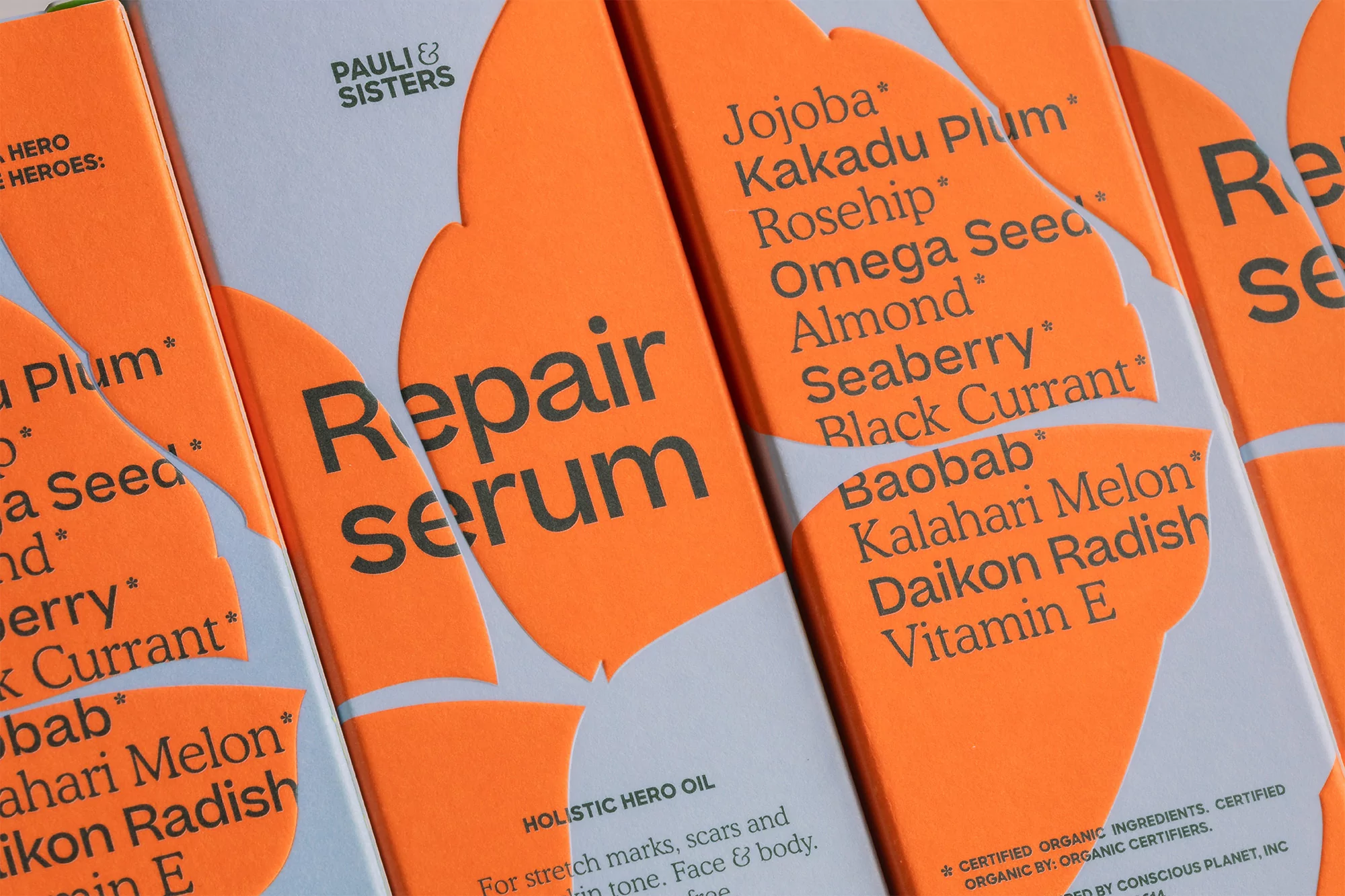 Close-up of several Pauli & Sisters 'Repair Serum' boxes with orange and white labels listing natural ingredients like Jojoba and Rosehip.