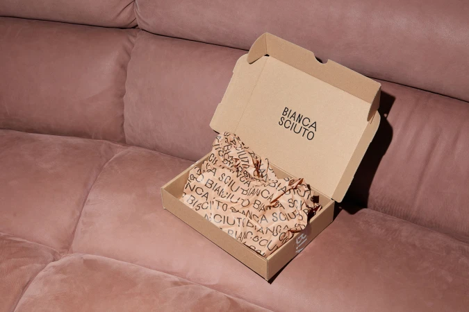 Open cardboard box with branded tissue paper, resting on a dusty rose velvet couch, in a studio shot with soft, indirect lighting.