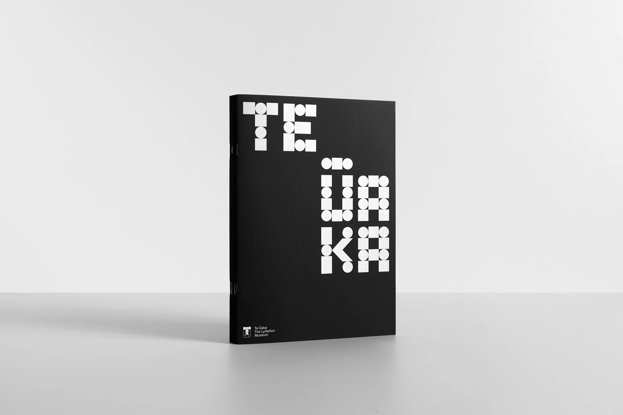 Black booklet with geometric white typography on a neutral white seamless studio background.
