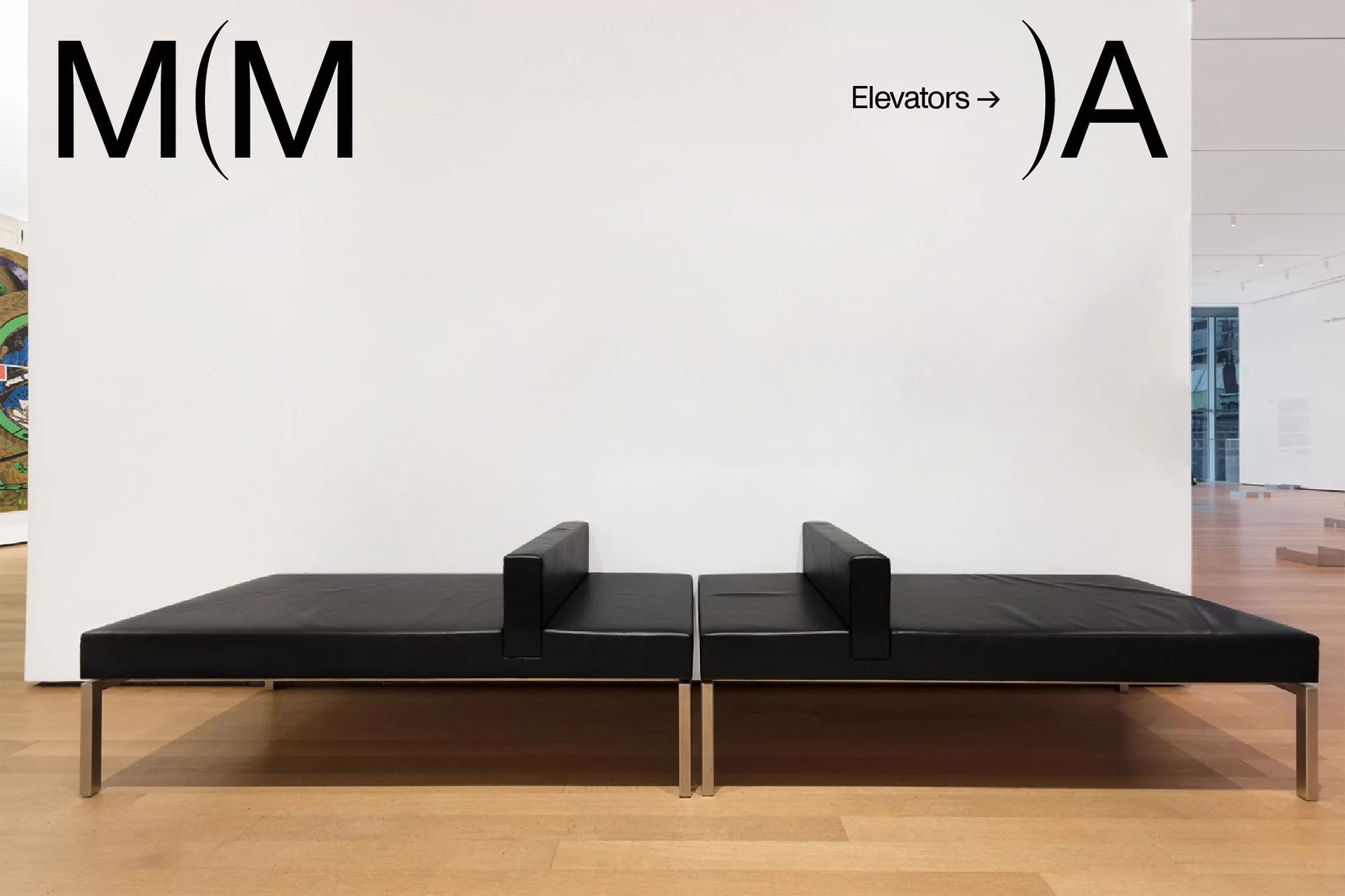 Interior view of a museum gallery with a modern black leather bench seat in front of a white wall with architectural signage.
