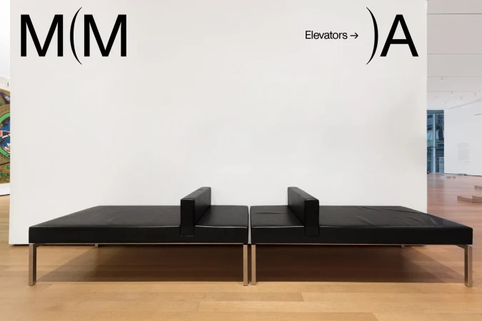 Interior view of a museum gallery with a modern black leather bench seat in front of a white wall with architectural signage.
